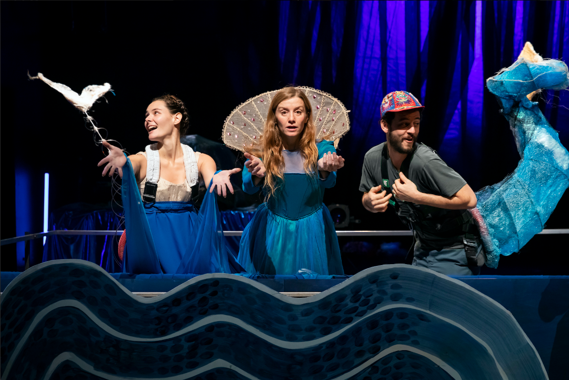 Photo du spectacle avec trois comédiens face au public (deux femmes et un hommes) devant un décors de mer