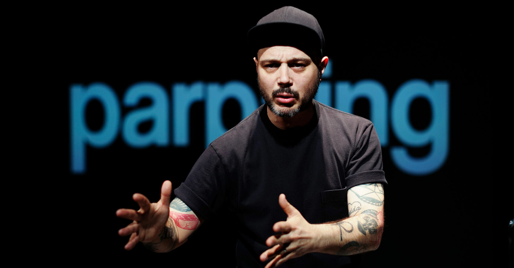 Photo de Nicolas Petisoff en train de parler avec un t-shirt et une casquette noir. Parpaing ecrit derrière lui