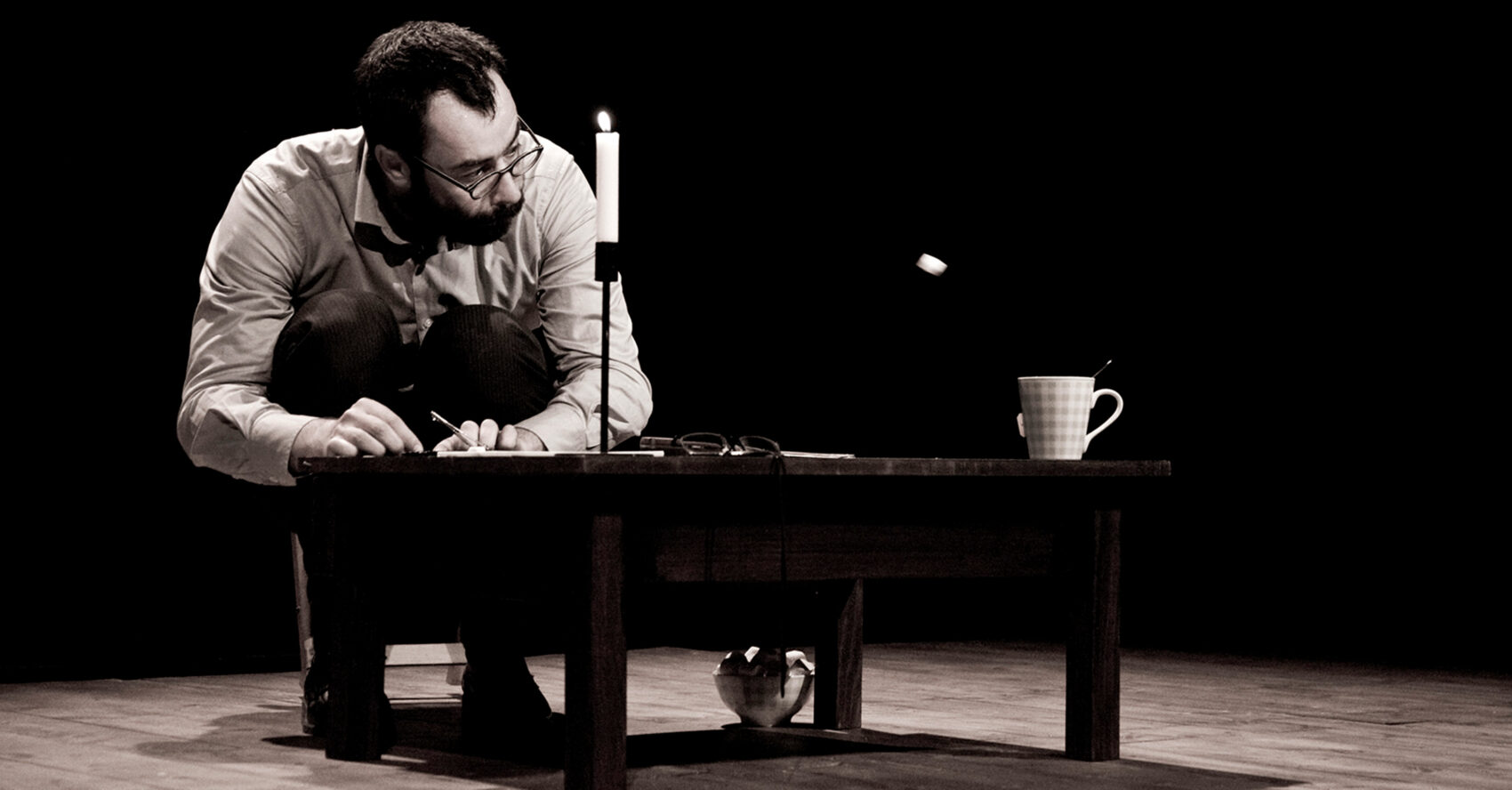 Un monsieur avec des lunettes est accroupi près d'une petite table. Sur la table, il a des objets comme une bougie et une tasse.