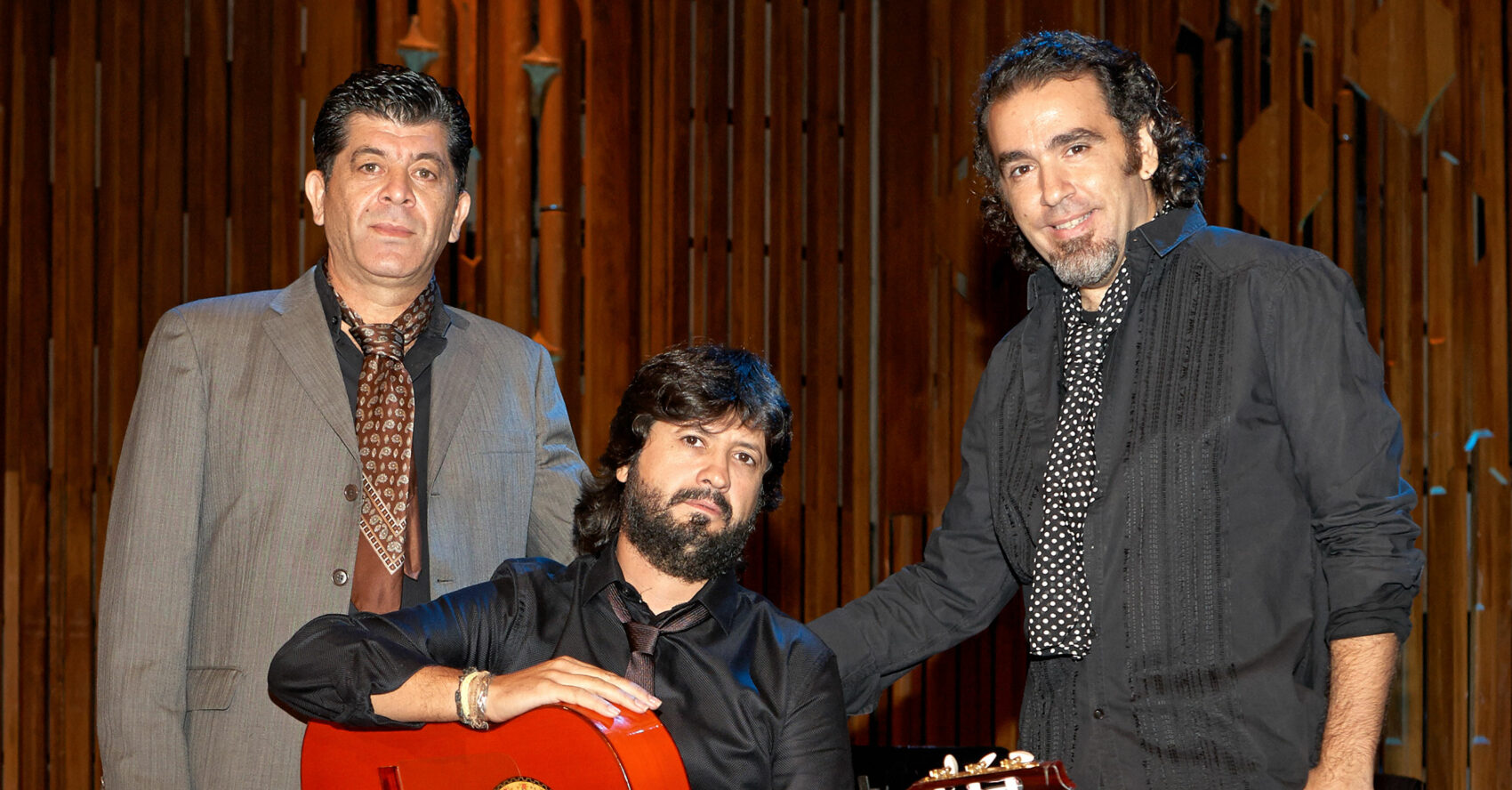 Deux musiciens sont debout autour d'un troisième qui est assis avec une guitare entre les mains.